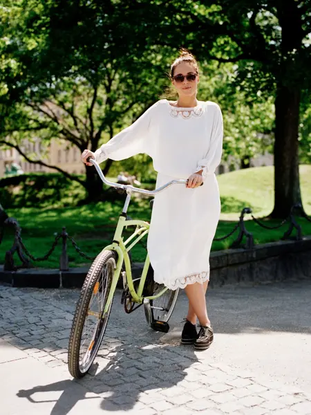 Dame med hvit kjole, brune sko og solbriller holder grønn sykkel med park i bakgrunnen 