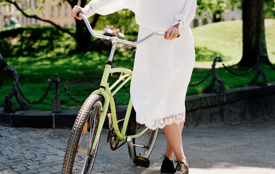 Dame med hvit kjole, brune sko og solbriller holder grønn sykkel med park i bakgrunnen 