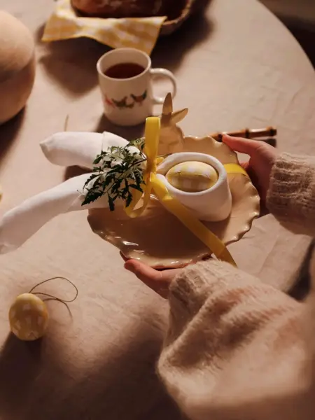 hender holder en tallerken med et egg og serviett dandert rundt, over et bord med duk og kaffekopp på.