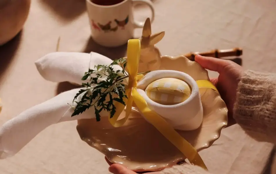 hender holder en tallerken med et egg og serviett dandert rundt, over et bord med duk og kaffekopp på.