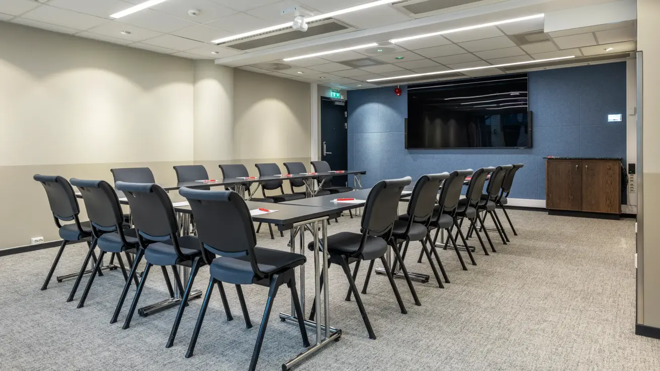 Møterom med u-bord oppsett, svarte stoler og bord, blå vegg i enden med sort TV skjerm