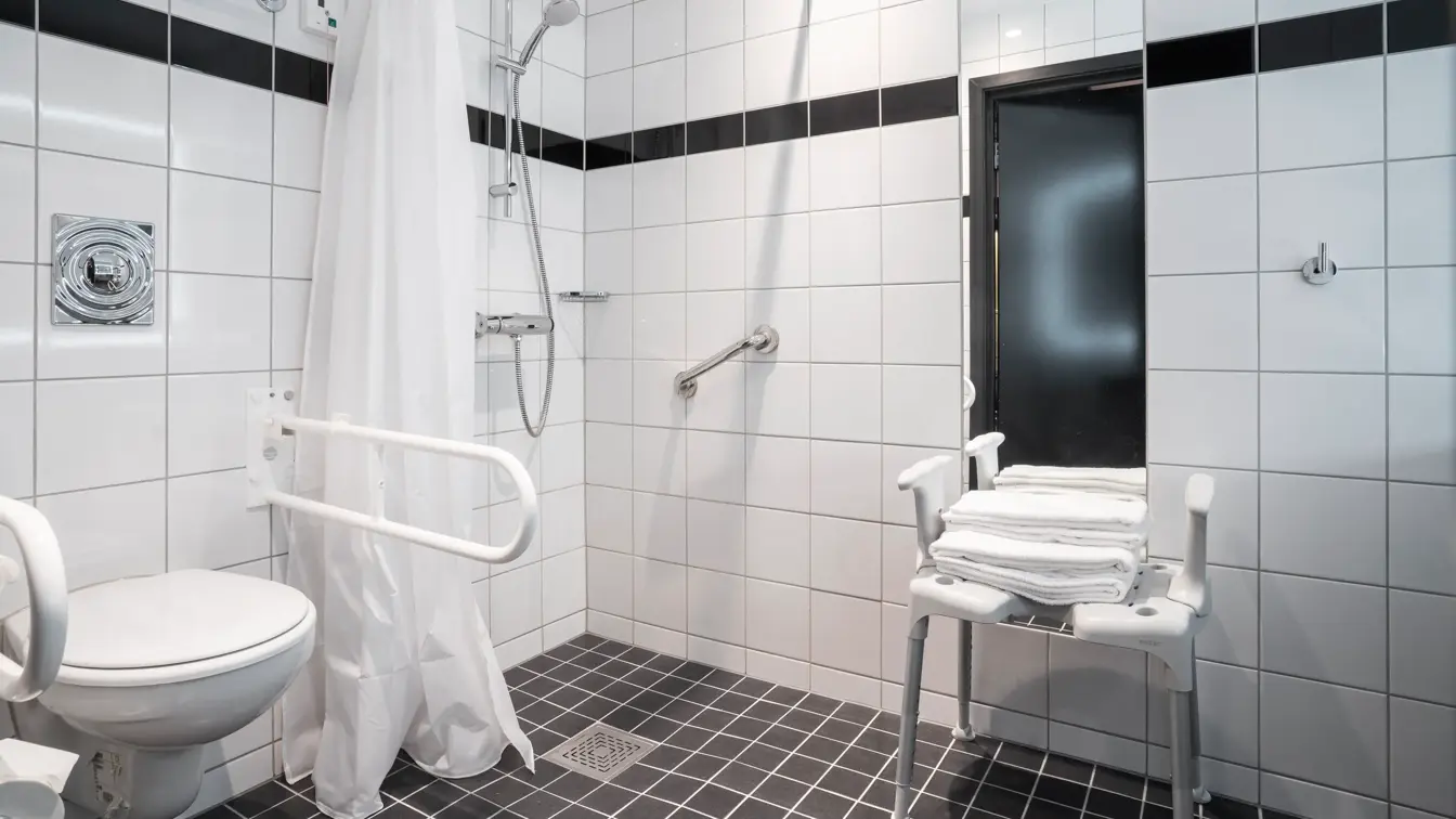 Dusjstol, dusj, toalett med støtte, hvite veggfliser, svarte gulvfliser. 