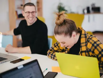 dame i oransje og svart rutete skjorte sitter og ler foran pc, mann i svart skjorte ler foran egen pc