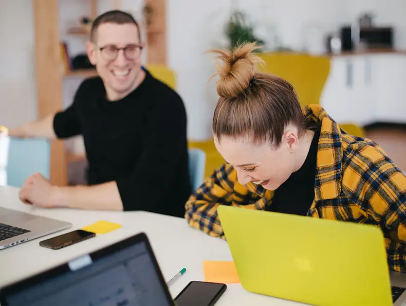 dame i oransje og svart rutete skjorte sitter og ler foran pc, mann i svart skjorte ler foran egen pc