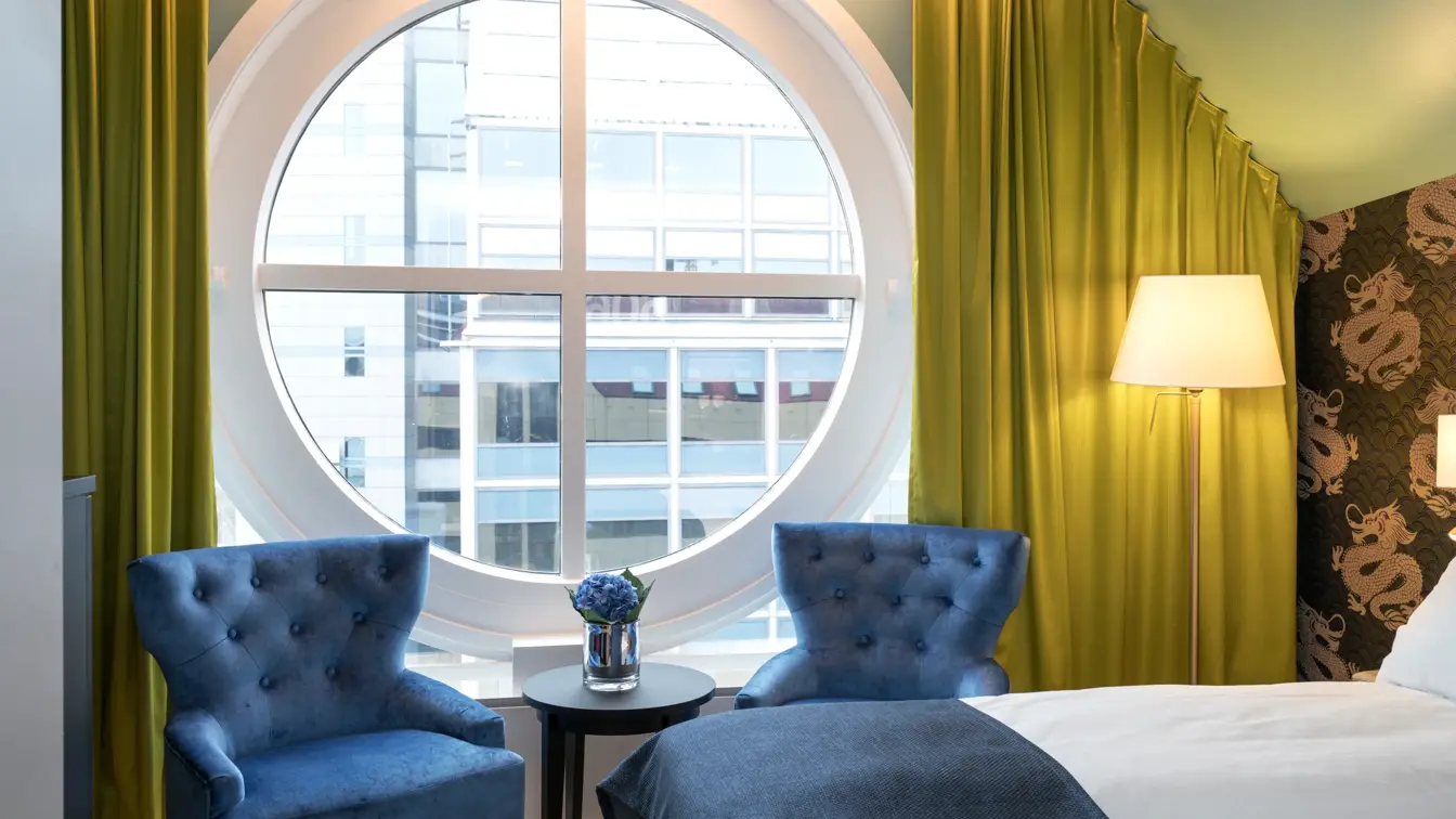 Rundt vindu med utsikt, dobbeltseng og blå lenestoler og gule gardiner på Thon hotel Terminus i Oslo sentrum nær Jernbanetorget