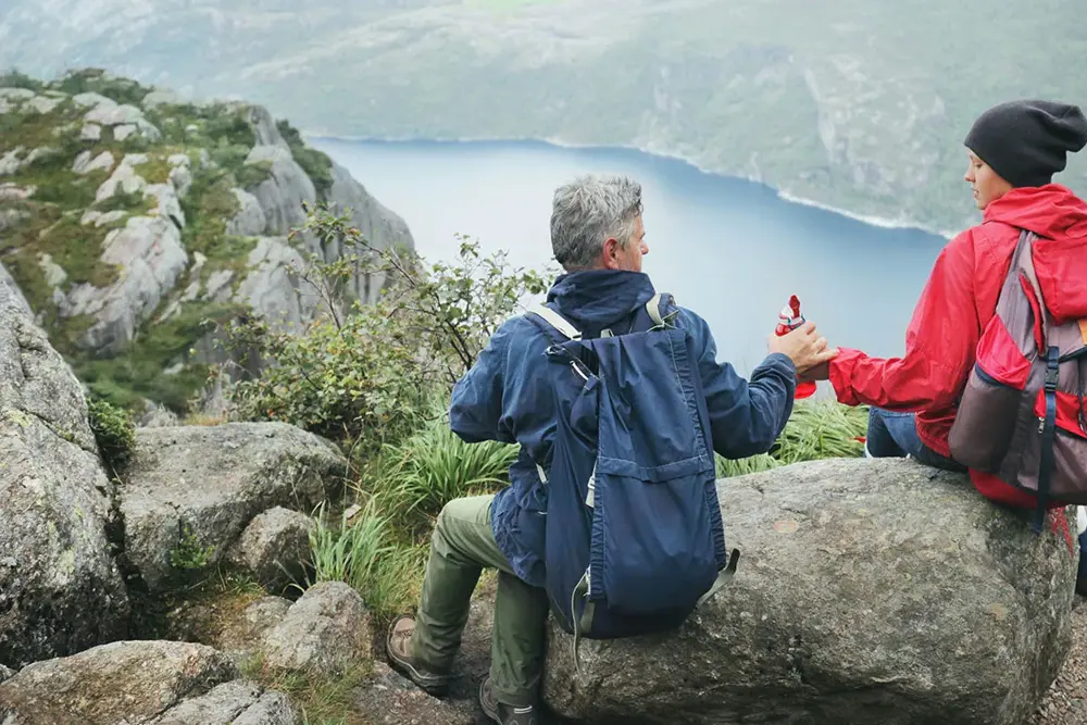 Jente og mann sitter med ryggen mot kamera på en stein høyt oppe på et fjell, med utsikt utover en fjord. Foto til artikkel om opplevelsesgaver til farsdag.