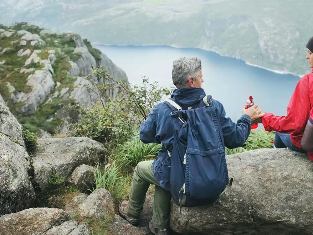 Jente og mann sitter med ryggen mot kamera på en stein høyt oppe på et fjell, med utsikt utover en fjord. Foto til artikkel om opplevelsesgaver til farsdag.