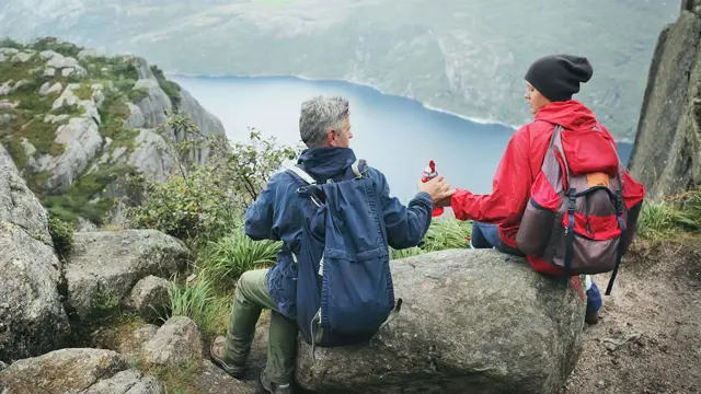 Jente og mann sitter med ryggen mot kamera på en stein høyt oppe på et fjell, med utsikt utover en fjord. Foto til artikkel om opplevelsesgaver til farsdag.