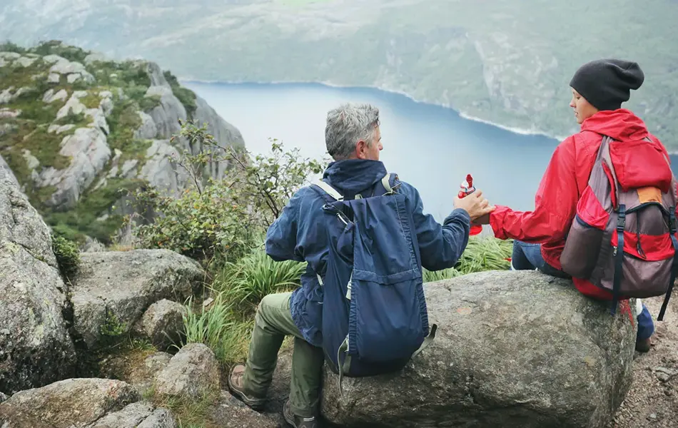 Jente og mann sitter med ryggen mot kamera på en stein høyt oppe på et fjell, med utsikt utover en fjord. Foto til artikkel om opplevelsesgaver til farsdag.