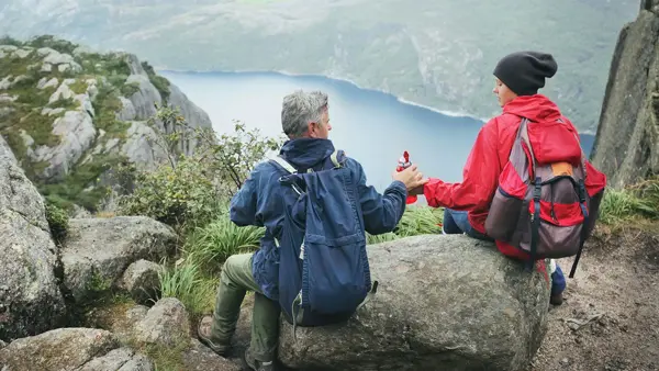 Jente og mann sitter med ryggen mot kamera på en stein høyt oppe på et fjell, med utsikt utover en fjord. Foto til artikkel om opplevelsesgaver til farsdag.