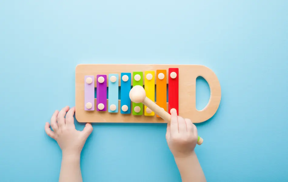 Baby hand holding hammer and playing colorful xylophone on light blue table background. Closeup. Music toy instrument of development for little kids. Point of view shot. Top down view.