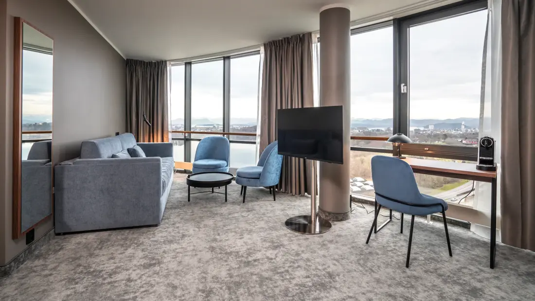 Suite med sofa og hvilestoler rundt avlasningsbord, skrivebord med kaffemaskin, lampe og tilhørende stol. TV, teppebelagt gulv. Høyt speil på veggen. Gulv-til-tak-vinduer langs hele veggen, med utsikt over byen.