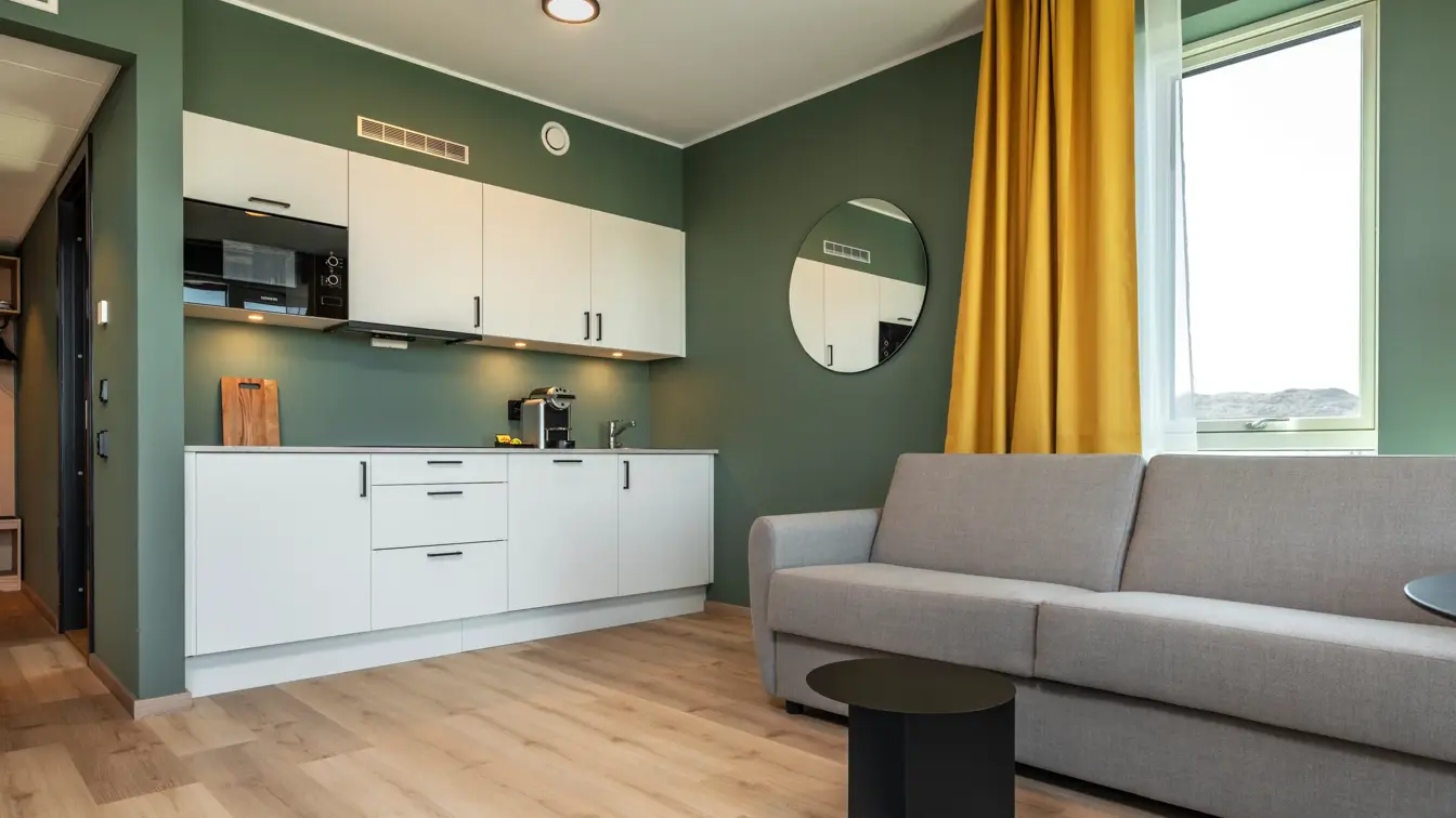 Leilighet med kjøkkenkrok, grå sofa, parkett, grønne vegger, vindu og gul gardin.
