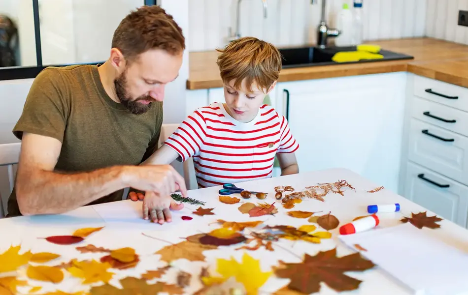 Far og sønn limer høstblader på papir. Foto.
