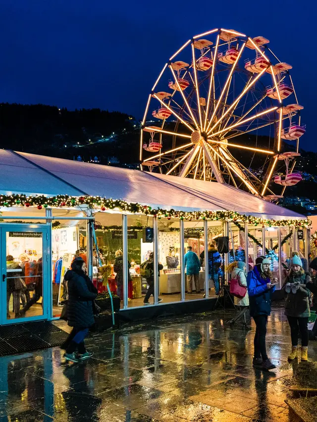 Folk rusler mellom opplyste telt og boder på julemarkedet i Bergen, med pariserhjul og bålpanne en våt vinterkveld.