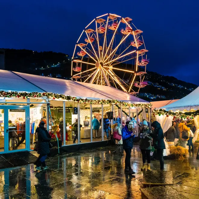 Folk rusler mellom opplyste telt og boder på julemarkedet i Bergen, med pariserhjul og bålpanne en våt vinterkveld.