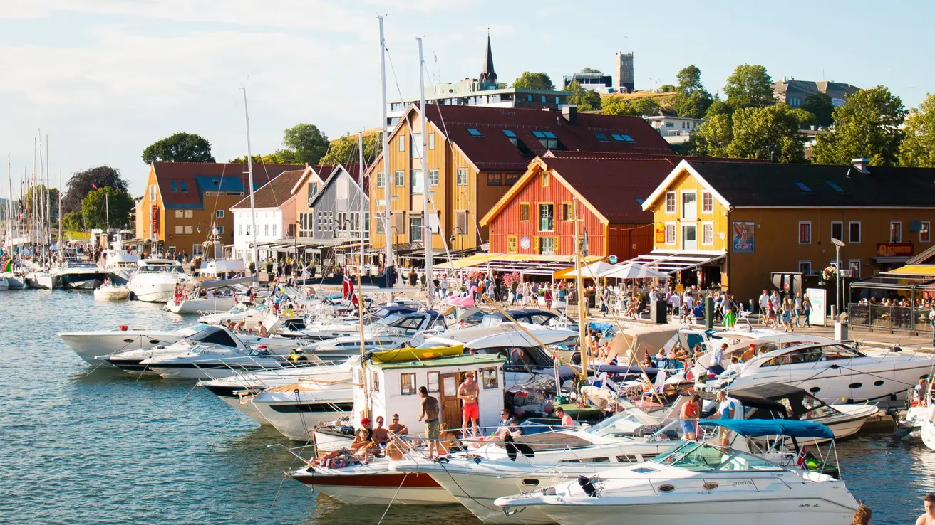 Brygga i Tønsberg en solskinnsdag på sommeren. Langs brygga ligger det båter og seilbåter i ulike størrelser, med mennesker, i flere rader. Til venstre ses Thon Hotel Tønsberg Brygge. Mot høyre ses rekke med restauranter. I bakgrunnen ses Slottsfjell