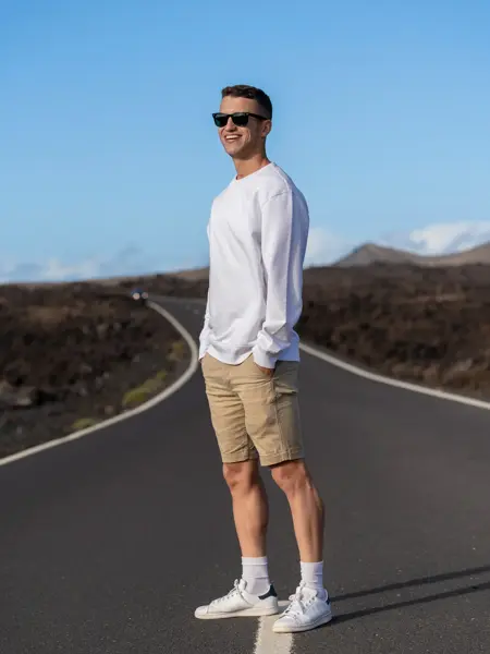 En mann i hvit genser, beige shorts, hvite sko og mørke solbriller står med hendene i lommene på en vei i et øde landskap. Mannen smiler mot kamera.