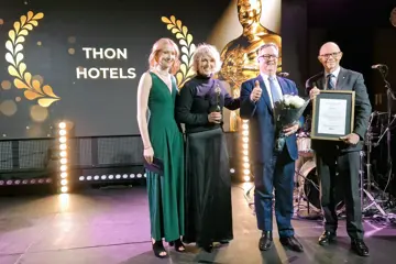To damer og to menn i pene klær står på scene under prisutdeling, med blomster og diplom, stor skjerm bak seg der det står Thon Hotels i gullskrift.