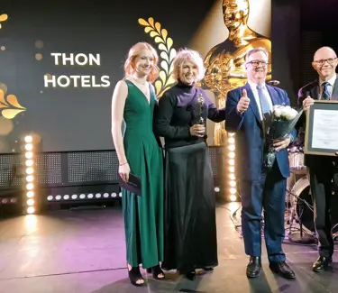 To damer og to menn i pene klær står på scene under prisutdeling, med blomster og diplom, stor skjerm bak seg der det står Thon Hotels i gullskrift.