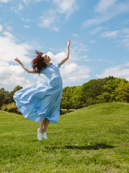Dame med lys blå kjole hopper på grønt gress med trær og blå himmel i bakgrunnen 