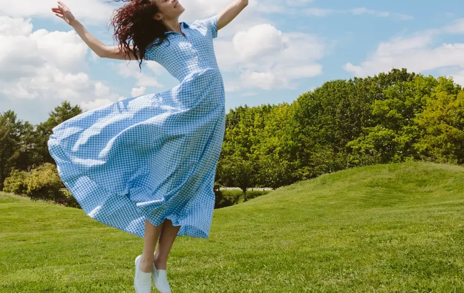 Dame med lys blå kjole hopper på grønt gress med trær og blå himmel i bakgrunnen 