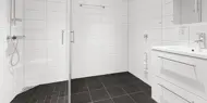 Tomt bad med hvit fliser på vegg og mørke fliser på gulv, dusjhjørne med glassvegger