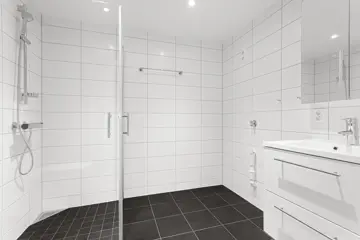 Tomt bad med hvit fliser på vegg og mørke fliser på gulv, dusjhjørne med glassvegger