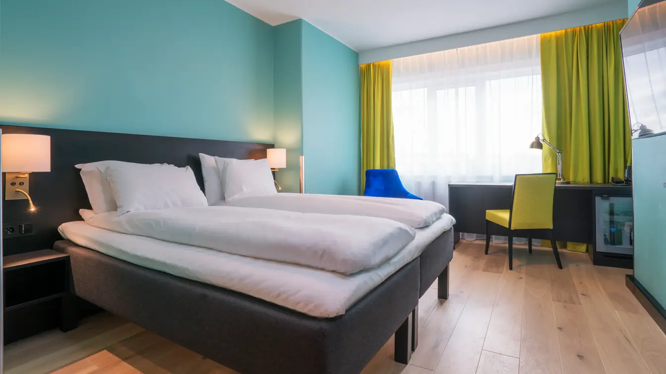 Twin room med hvit sengetøy, gule gardiner, skrivepult og blå lenestol på Thon Hotel Europa i Oslo sentrum rett ved Slottsparken