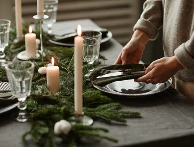 Side view close up of unrecognizable woman setting up dining table decorated for Christmas with fir branches and candles in grey tones, copy space