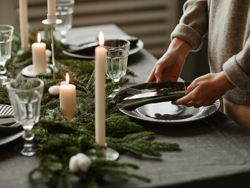 Side view close up of unrecognizable woman setting up dining table decorated for Christmas with fir branches and candles in grey tones, copy space