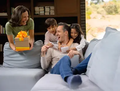 Mann sitter i sofa og får gave av kvinne og to barn, alle smiler. Foto til artikkel om gaver til pappatyper på farsdag. 