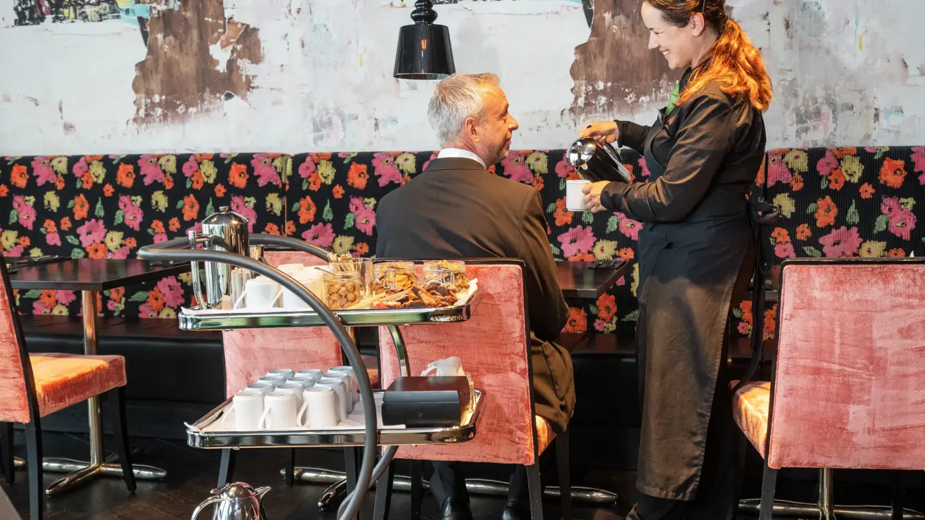 Servitør heller kaffe i en kaffekopp til en gjest, tapetvegg med mønster, spisebord i bakgrunn. 