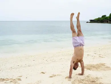 Mann står på henda på øde strand i lilla badebukse.