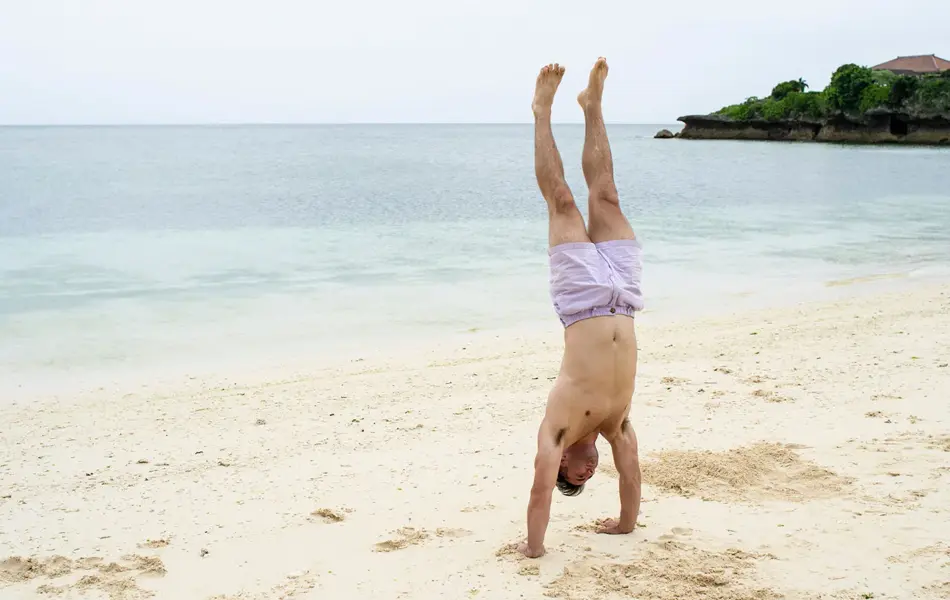 Mann står på henda på øde strand i lilla badebukse.