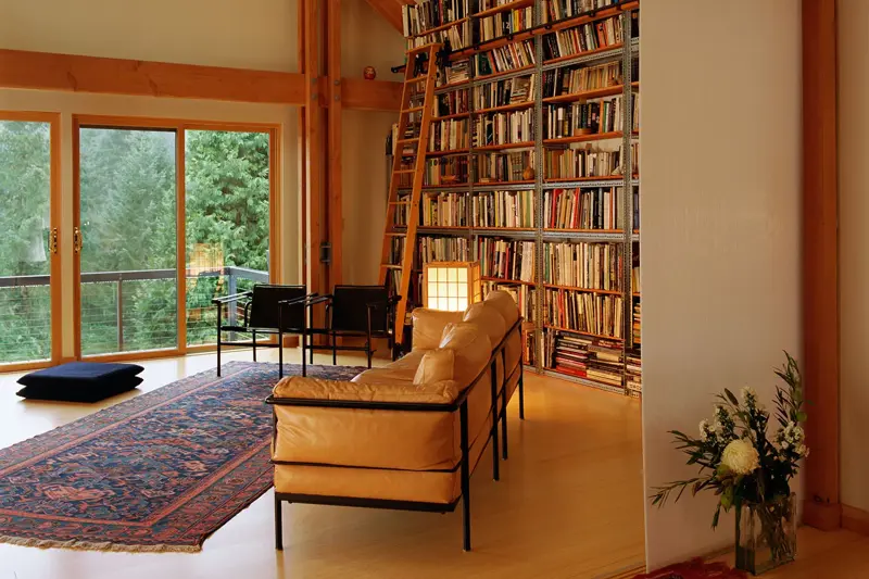 Et rom med høyt under taket og en høy bokhylle med en stige lent inntil. I forgrunnen står en sofa og et teppe på gulvet.
