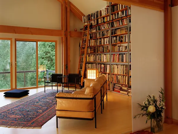 Et rom med høyt under taket og en høy bokhylle med en stige lent inntil. I forgrunnen står en sofa og et teppe på gulvet.