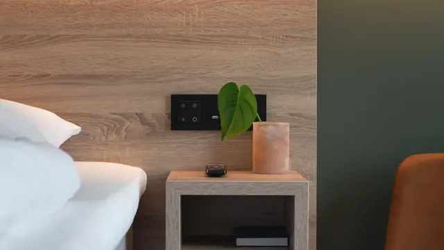 Detaljbilde av sengegavl i lyst træ, lite vegghengt sengbord. Mørkegrønn tapet og en stol i lysebrunt skinn med sorte ben.  I standard dobbelt rom på Thon Hotel Norge i Kristiansand.