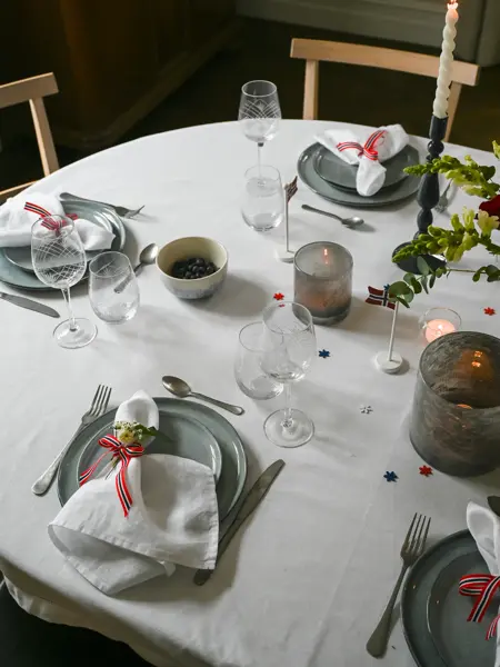 Ovalt bord dekket med hvit duk, grått service, hvite linservietter, telysholdere og grønne grener.