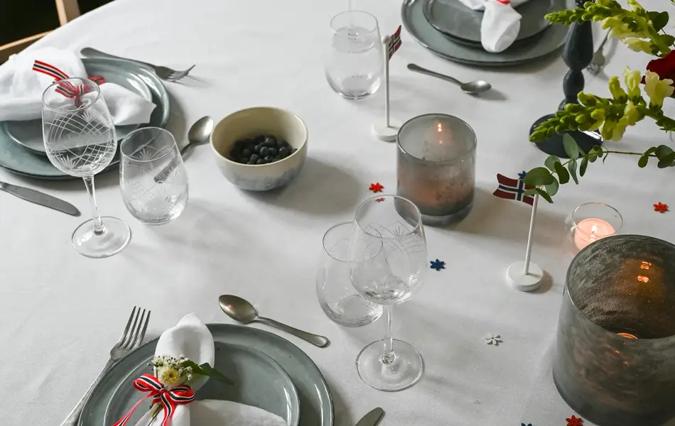 Ovalt bord dekket med hvit duk, grått service, hvite linservietter, telysholdere og grønne grener.