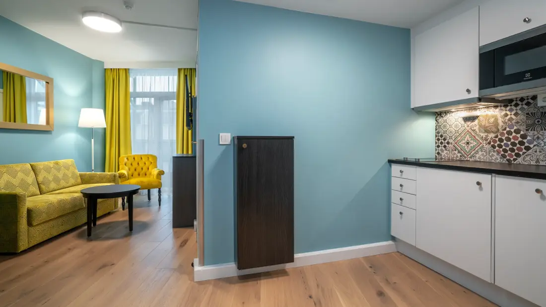 Lite kjøkken, mikrobølgeovn, gul sofa, gul stol, gule gardiner, vindu fra gulv til tak, blå vegger, salongbord, parkettgulv. 