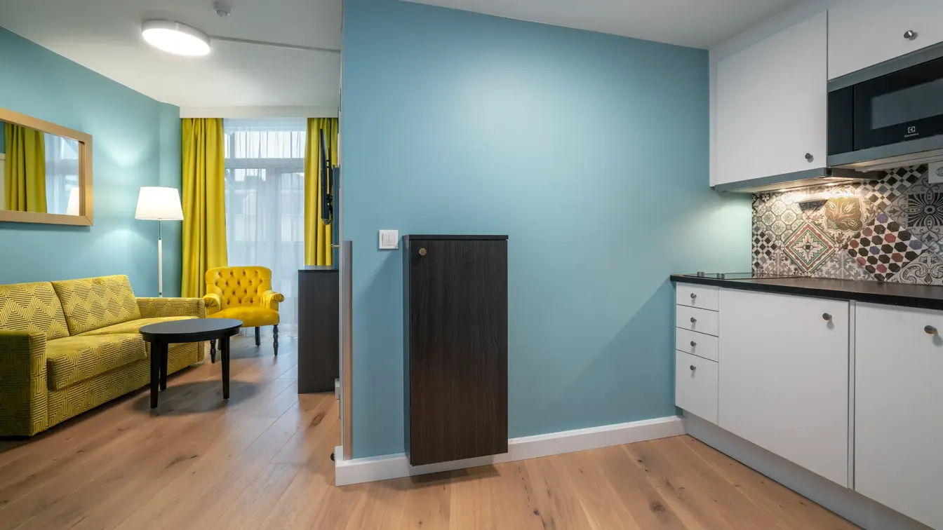 Lite kjøkken, mikrobølgeovn, gul sofa, gul stol, gule gardiner, vindu fra gulv til tak, blå vegger, salongbord, parkettgulv. 