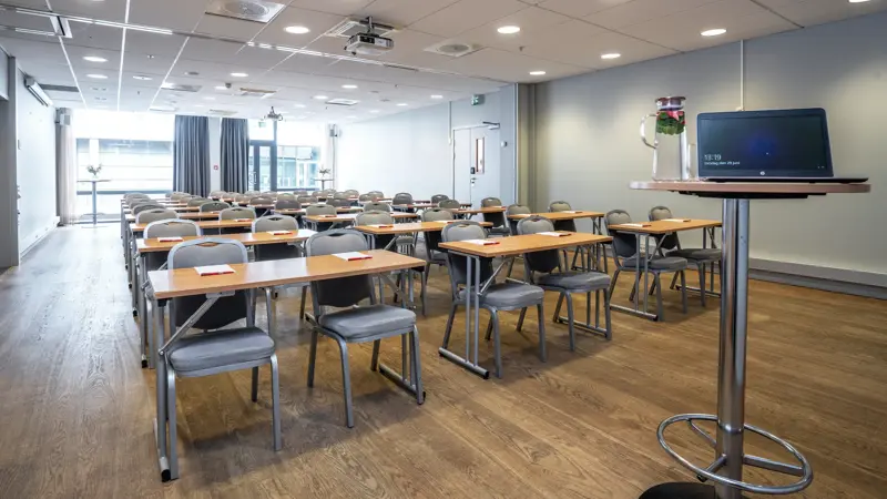 Konferanserom sett forfra. Høyt, rundt bord med PC, bord og stoler i klasseromsoppsett. Vinduer bakerst. Parkettgulv.