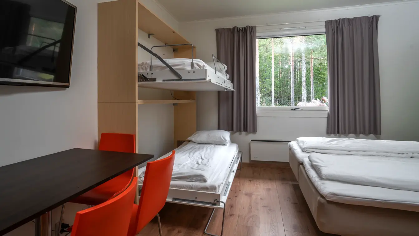 Studioleilighet med dobbeltseng og køyeseng, vindu med grå gardiner, skrivebord, rød stoler og TV på veggen.