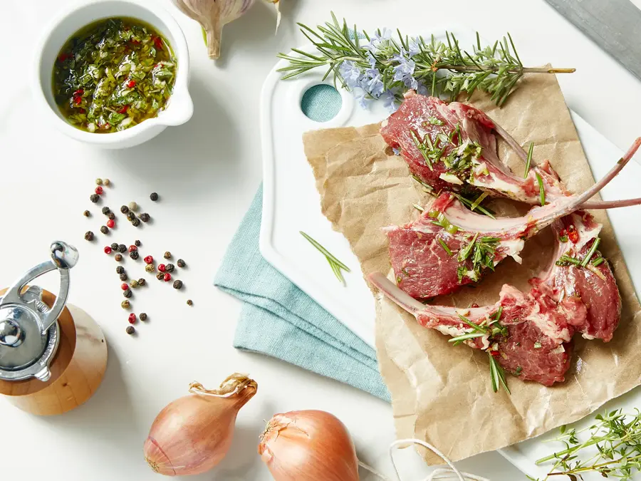 overhead view of raw fresh lamb chops on a cutting board with gremolata, rosemary, shallots, garlic, thyme, peppercorns and parsey