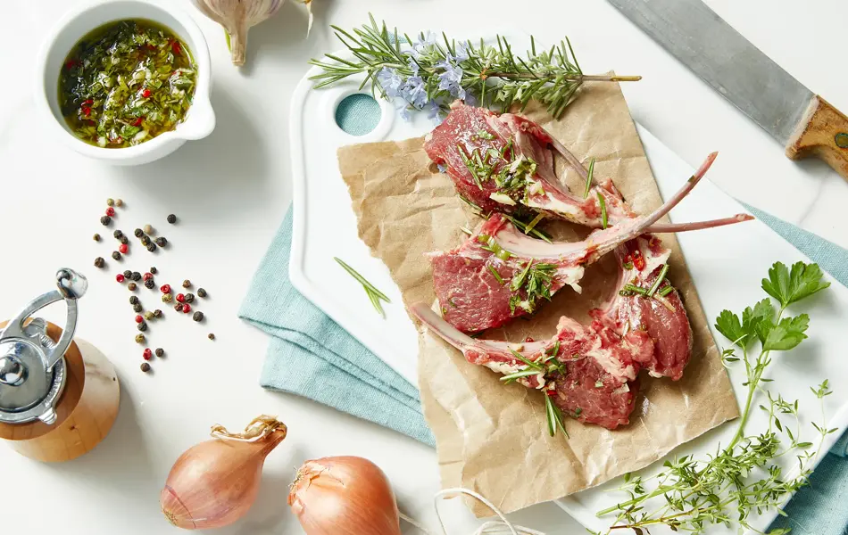 overhead view of raw fresh lamb chops on a cutting board with gremolata, rosemary, shallots, garlic, thyme, peppercorns and parsey