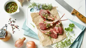 overhead view of raw fresh lamb chops on a cutting board with gremolata, rosemary, shallots, garlic, thyme, peppercorns and parsey