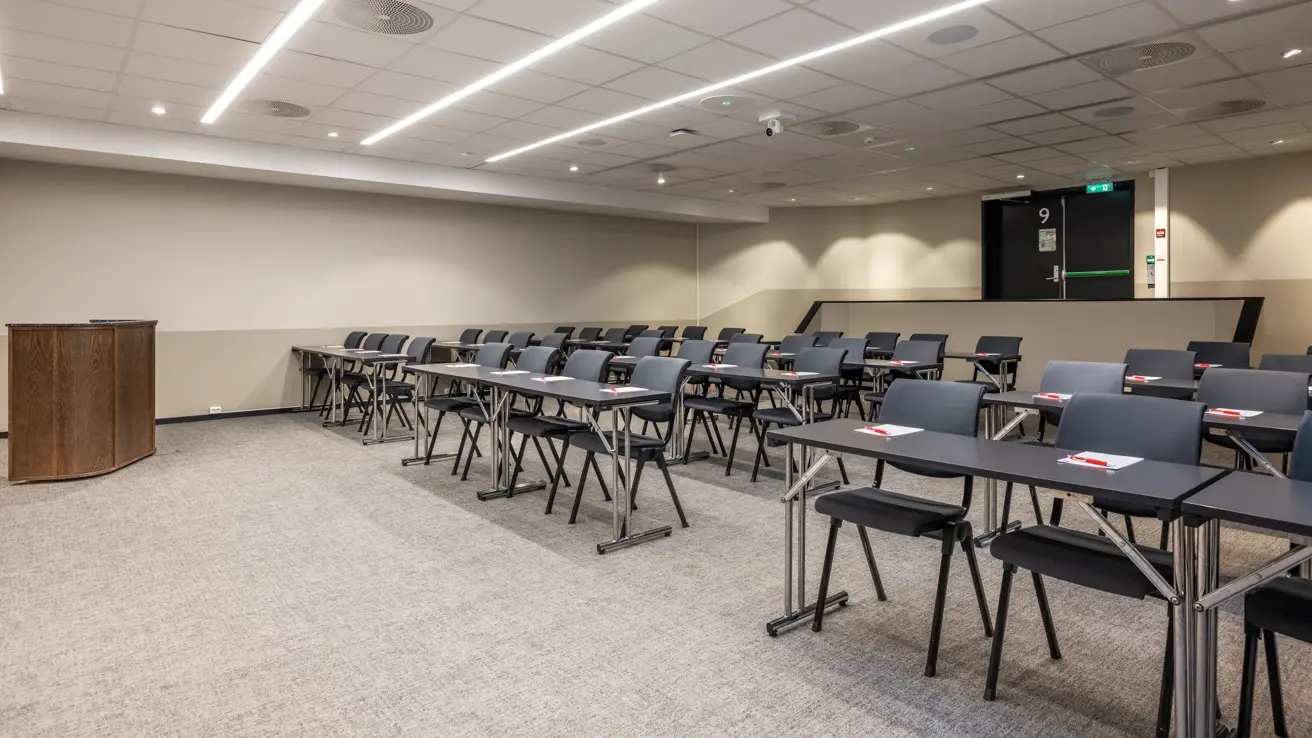 Møterom med klasseroms oppsett, talerstol foran, grått gulvteppe, trapp opp bakerst til inngangsdør.