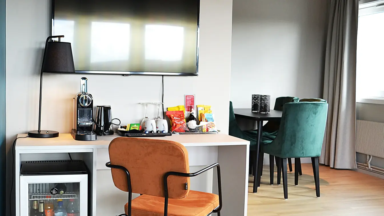 Tv, minibar, skrivebord, stoler, lampe, rundt bord med stoler, grå gardiner, vindu