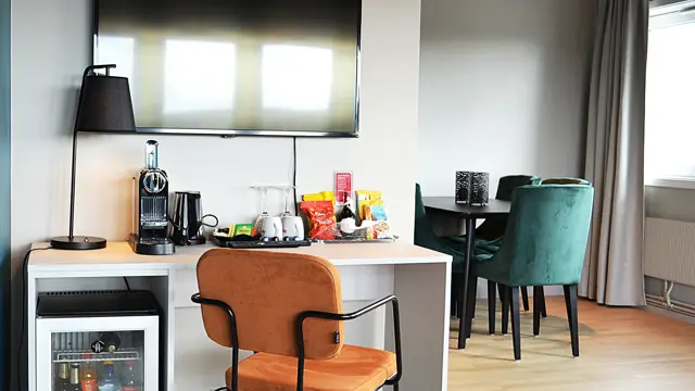 Tv, minibar, skrivebord, stoler, lampe, rundt bord med stoler, grå gardiner, vindu
